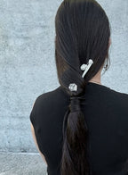 A person viewed from behind with long dark brown hair styled in a low ponytail, secured with two silver hair accessories against a textured gray wall.