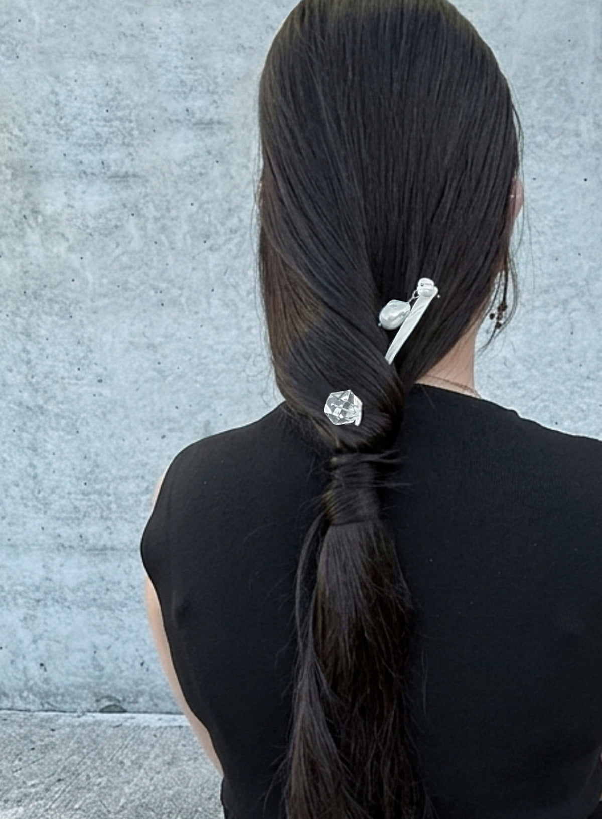 A person viewed from behind with long dark brown hair styled in a low ponytail, secured with two silver hair accessories against a textured gray wall.