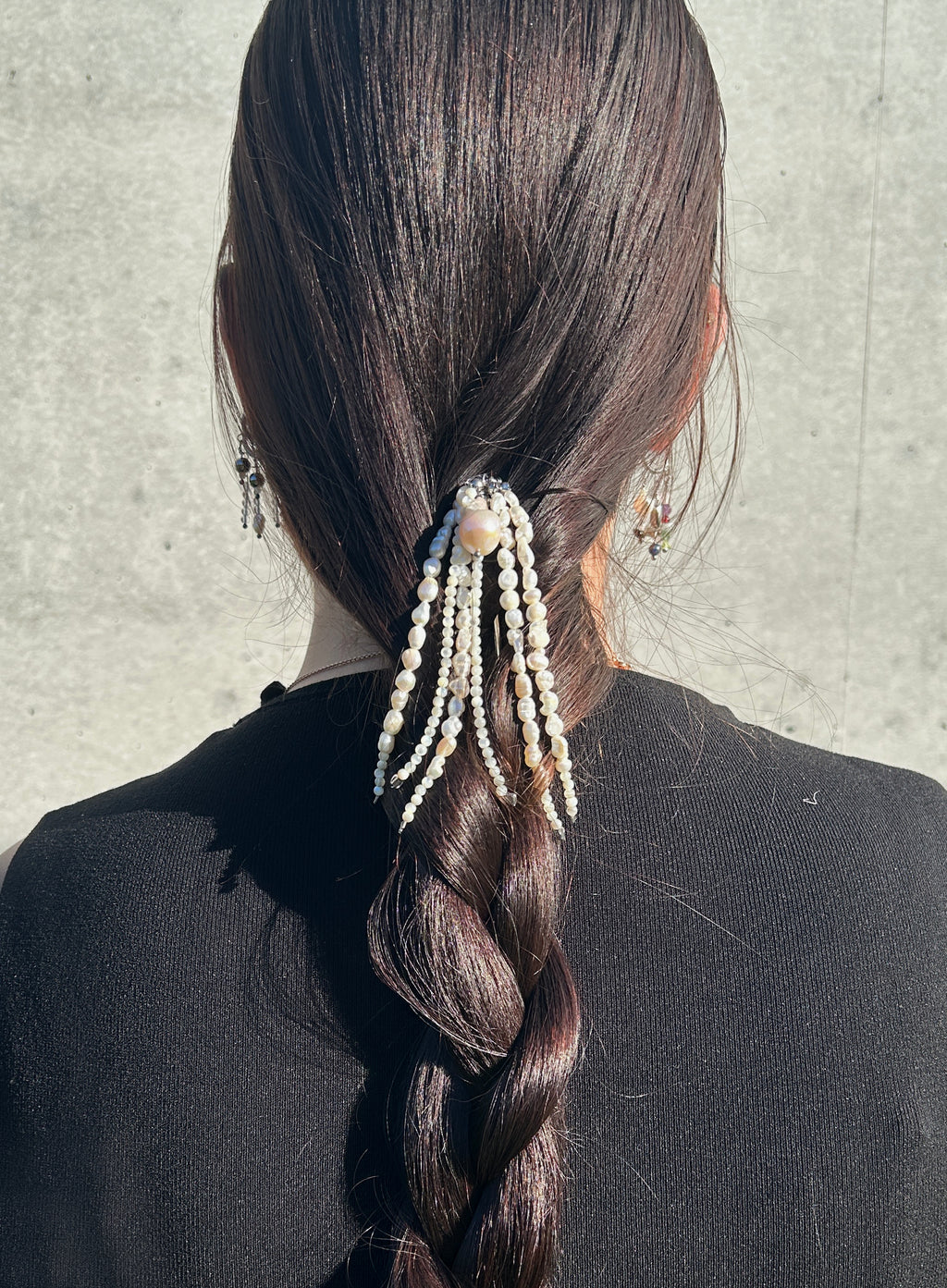 A dark brown beaded hair accessory with pearl-like beads and dangling strands, styled in a low ponytail on a person with long dark hair, shown from the back against a light gray concrete wall.