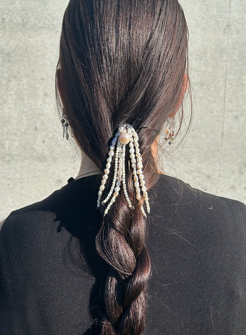 A dark brown beaded hair accessory with pearl-like beads and dangling strands, styled in a low ponytail on a person with long dark hair, shown from the back against a light gray concrete wall.