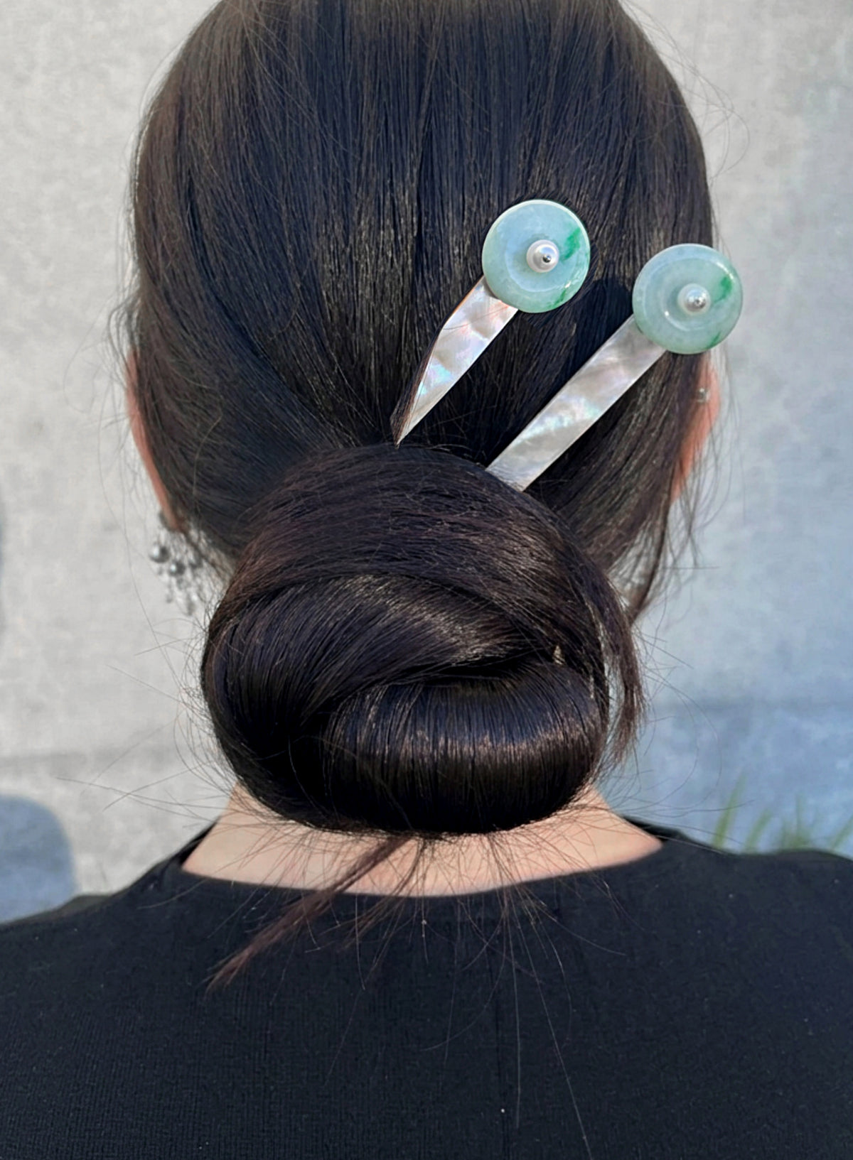 A person viewed from the back wearing a dark bun hairstyle, styled with two hairpins featuring round light green stone tops and iridescent white mother-of-pearl handles, set against a neutral gray wall.