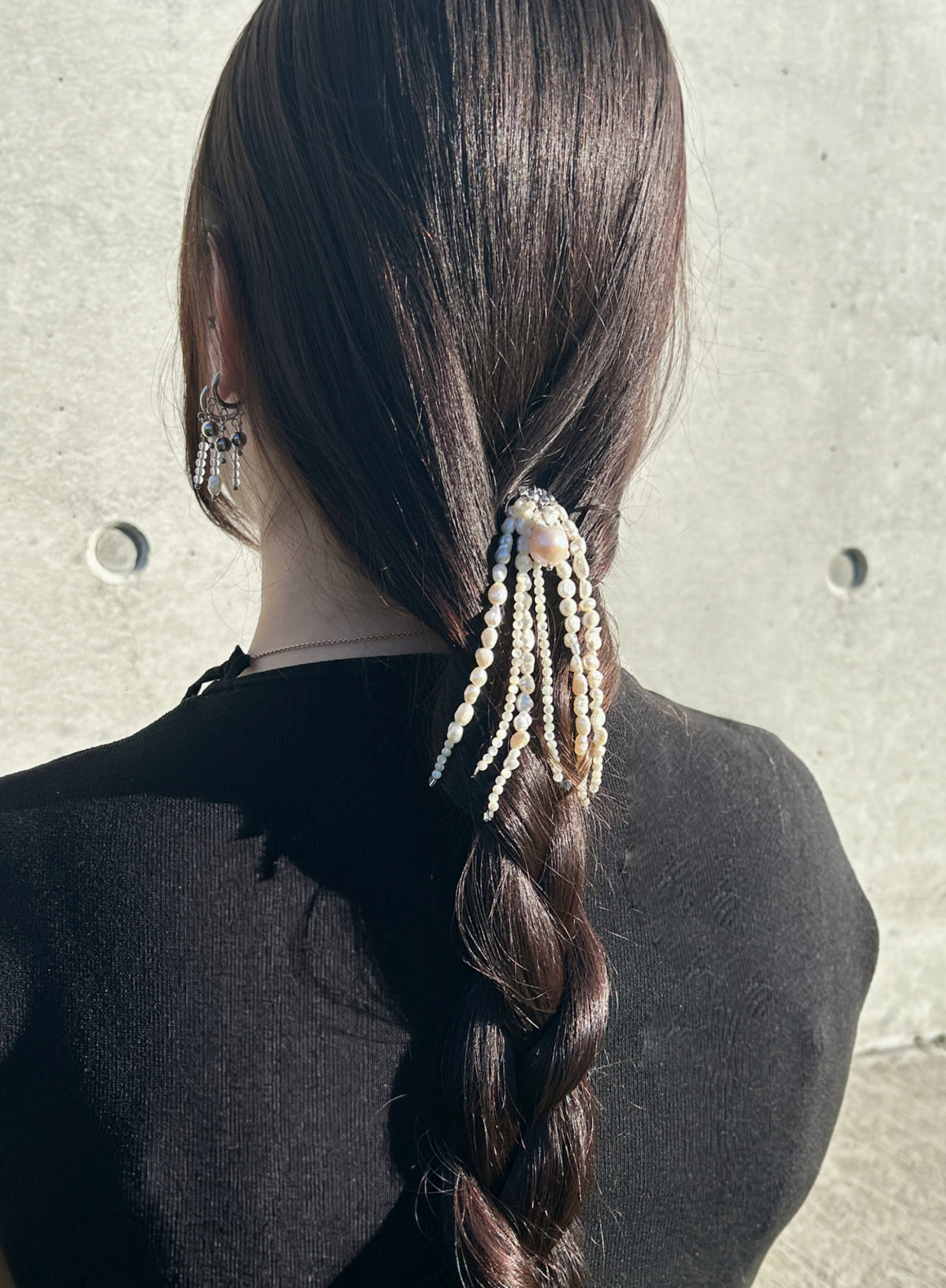A woman viewed from behind wearing a long dark brown braided ponytail adorned with dangling cream pearl-like strands, set against a light gray concrete wall.