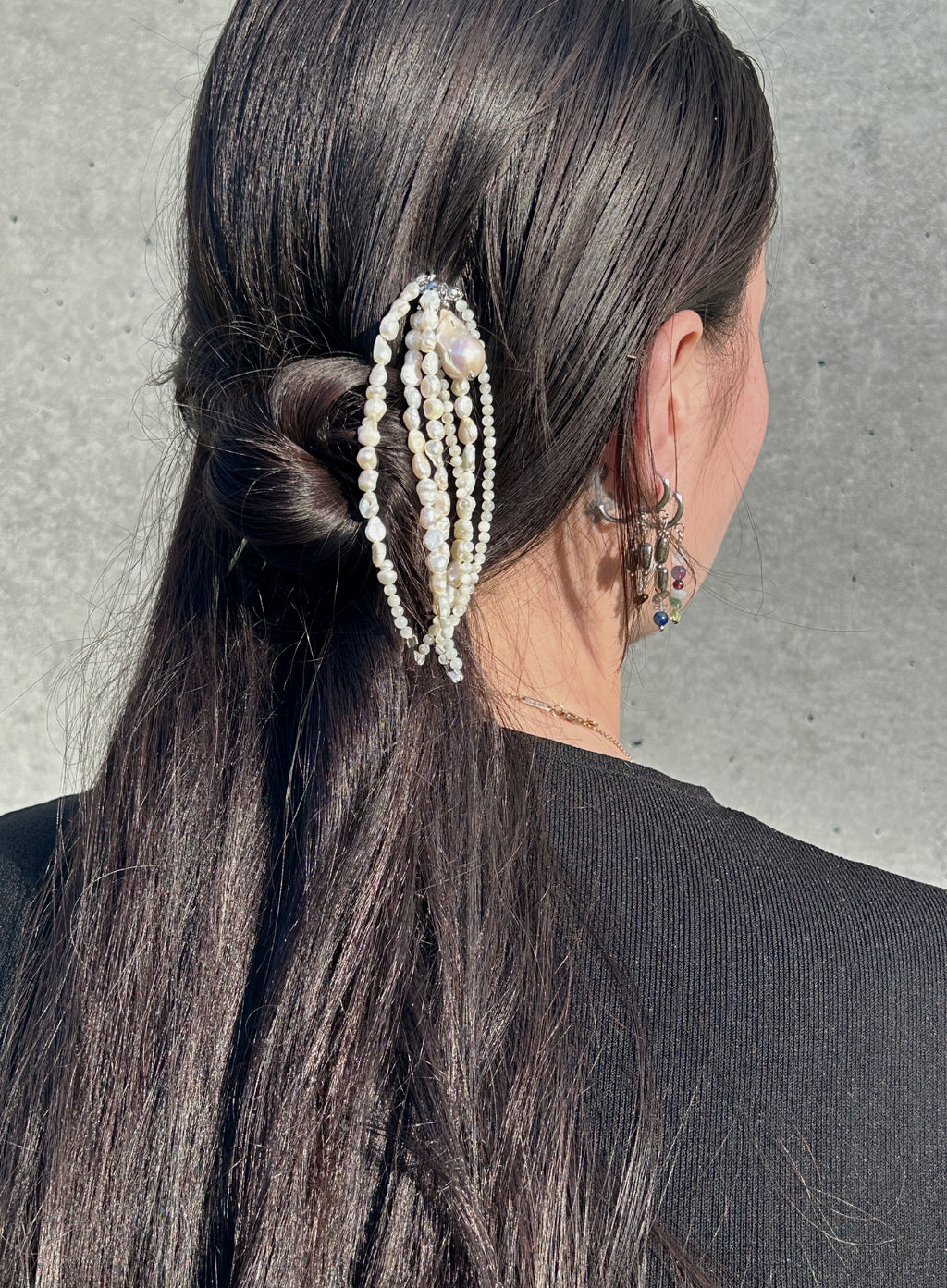 A close-up back view of a person with long dark hair wearing a decorative hair accessory made of multiple strands of white faux pearls with small bead details, against a gray concrete wall.
