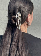 A close-up back view of a person with long dark hair wearing a decorative hair accessory made of multiple strands of white faux pearls with small bead details, against a gray concrete wall.
