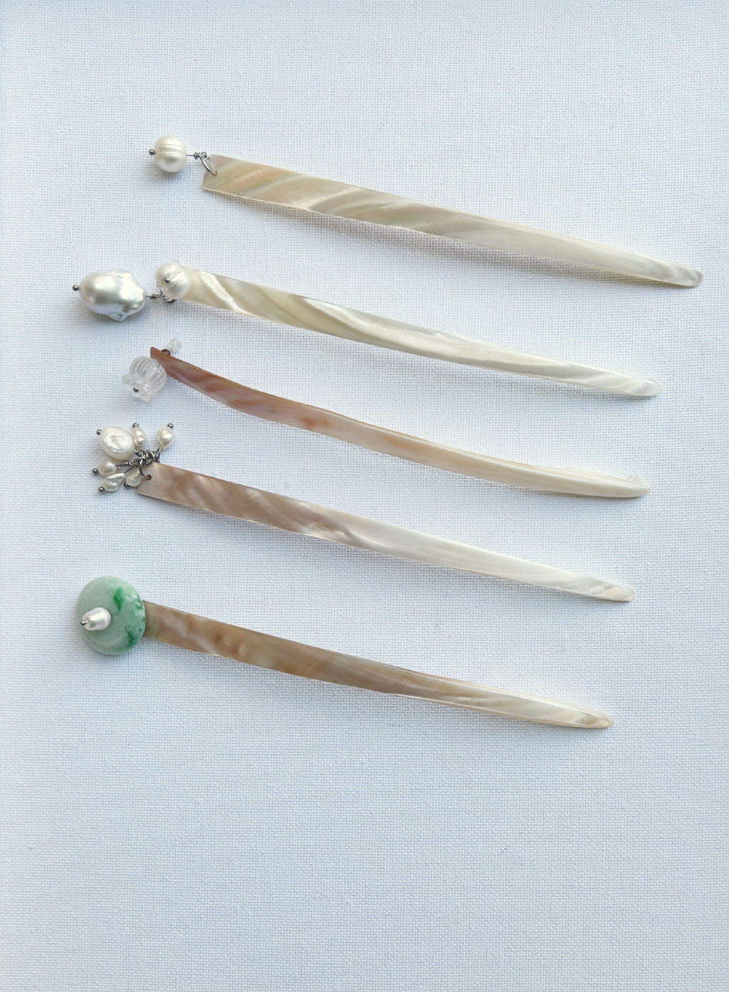 Five long hair sticks or hair pins made of iridescent mother-of-pearl with various bead and gemstone accents, arranged diagonally on a light blue background.