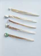 Five long hair sticks or hair pins made of iridescent mother-of-pearl with various bead and gemstone accents, arranged diagonally on a light blue background.