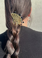 A close-up back view of a person wearing a brownfan-shaped hair pin with small black pearls, holding a dark brown braided ponytail. The person is wearing a black top against a neutral concrete wall.