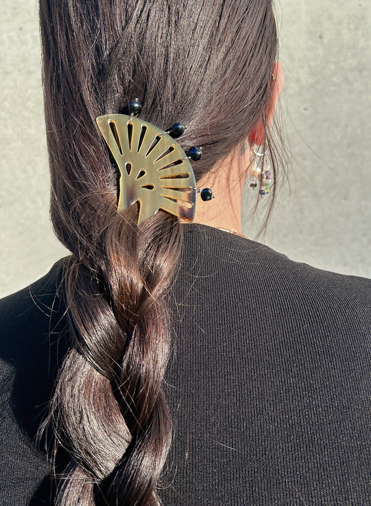 A close-up back view of a person wearing a brownfan-shaped hair pin with small black pearls, holding a dark brown braided ponytail. The person is wearing a black top against a neutral concrete wall.
