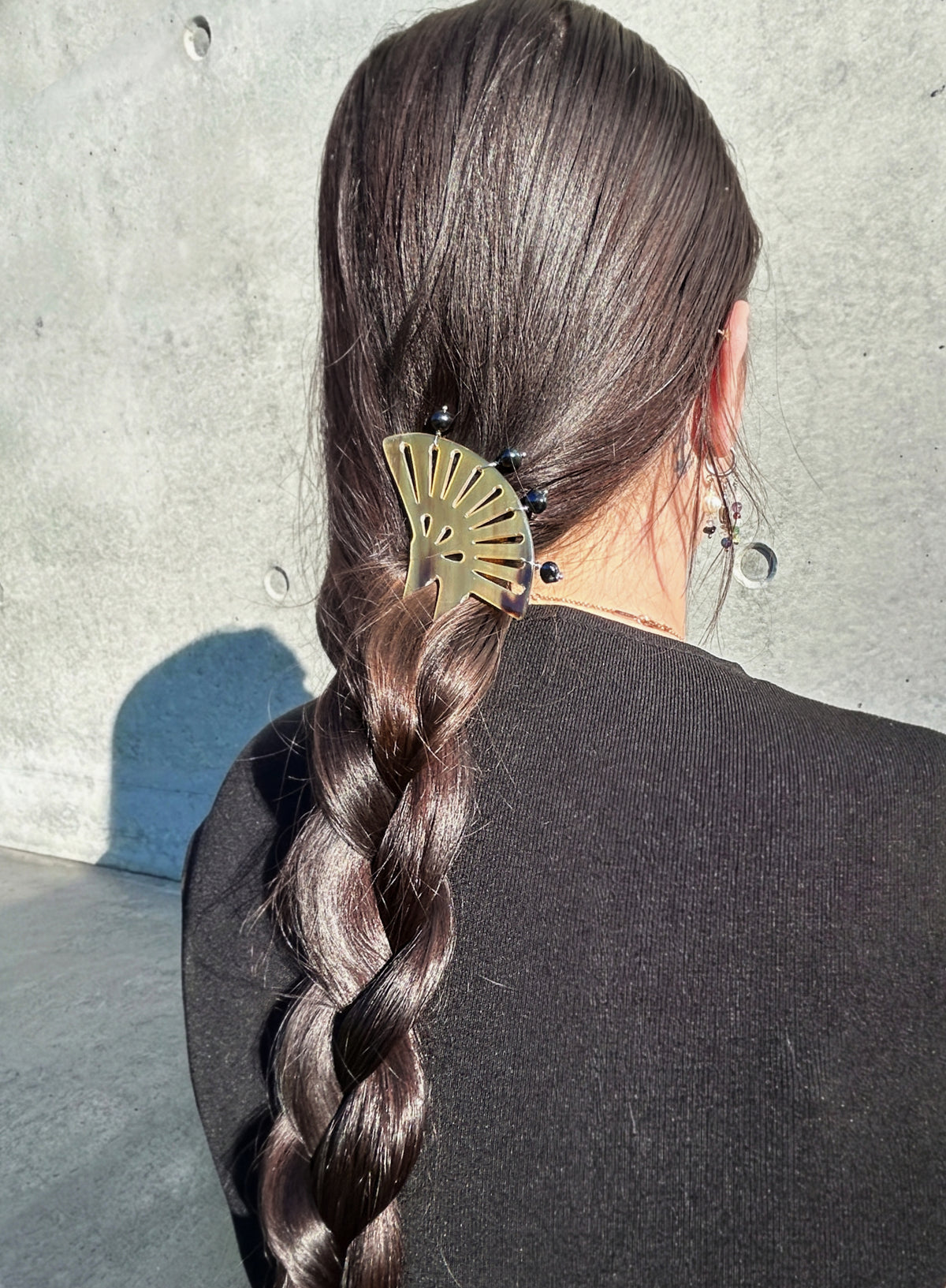 A person viewed from behind wearing a dark brown braided hair accessory with a brown fan-shaped hair pin against a gray concrete wall.