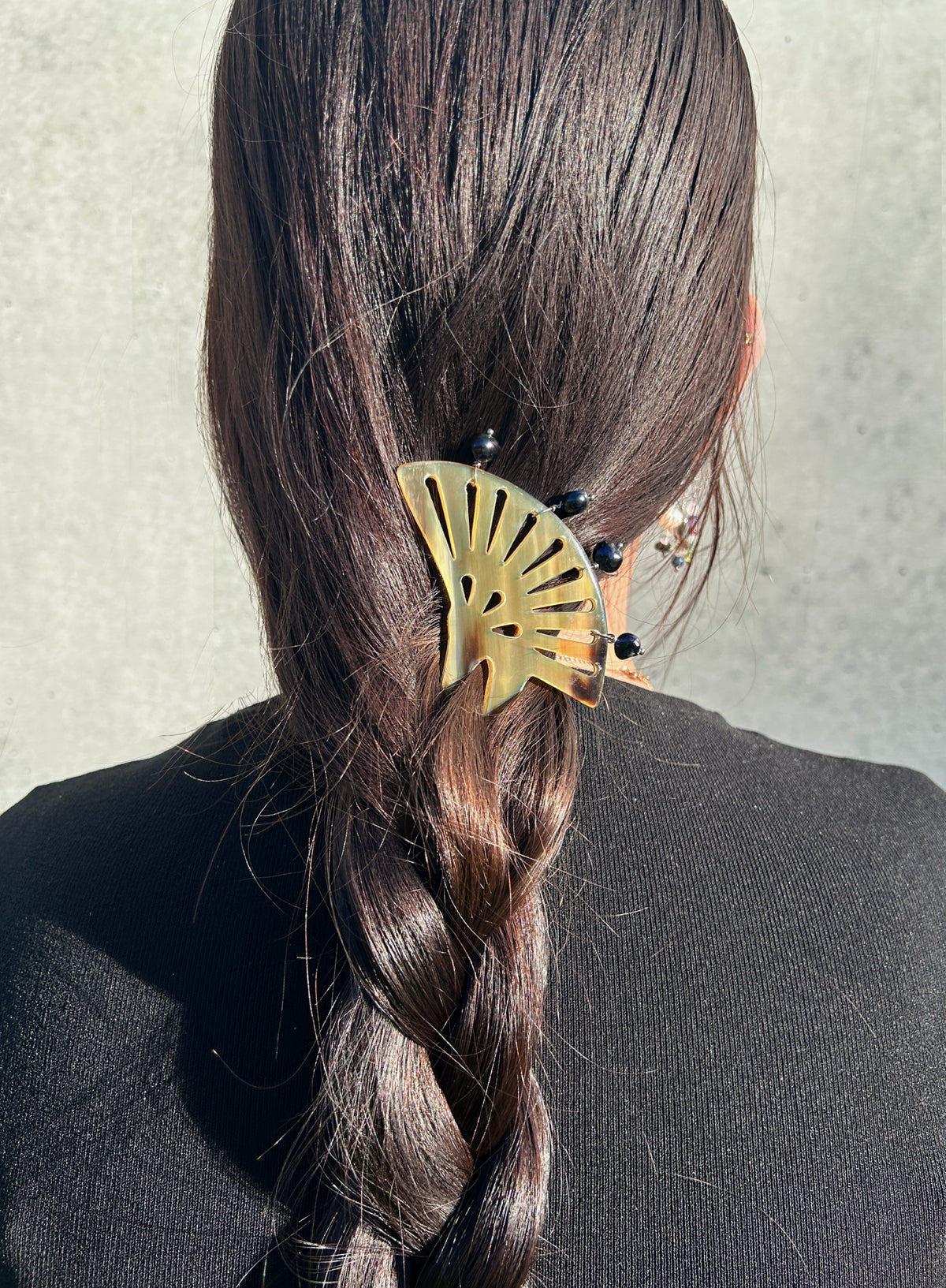 A close-up back view of a person with long dark brown hair in a braid, wearing a brown fan-shaped hair accessory with small black pearls.