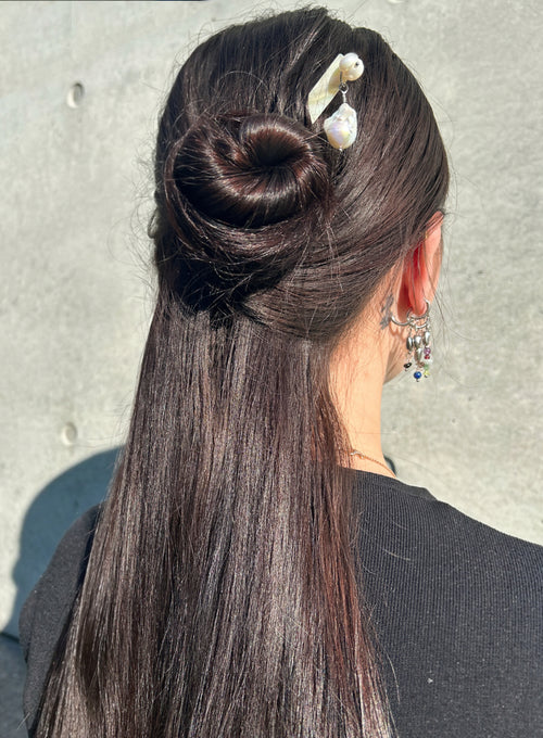 A woman shown from the back wearing a dark brown half-up bun with a pearl hair pin and long, straight dark brown hair. She is wearing a black top against a gray concrete wall.