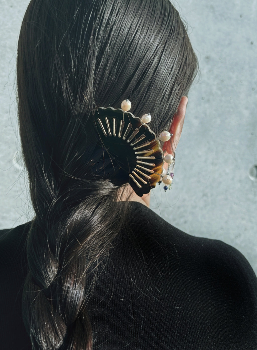 A close-up back view of a person with long dark hair wearing a black hair accessory with gold-toned fan-like detailing and pearl-like beads, against a textured light gray background.