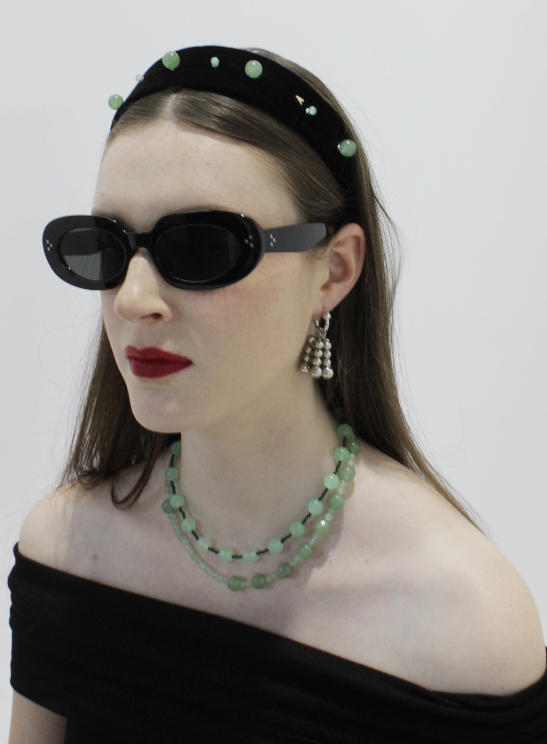Woman wearing black sunglasses, a black headband with green beads, and a black off-shoulder top against a white background.
