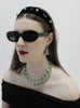 Woman wearing black sunglasses, a black headband with green beads, and a black off-shoulder top against a white background.