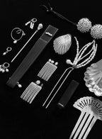 A monochrome flat-lay arrangement of fashion accessories on a black background, including multiple pearl-like hair combs, dangling earrings, a long comb, a fan-shaped hair tool, a shell-shaped hair piece, beaded strands, textured spherical ornaments, and a small black rectangular item with visible CHANEL branding.