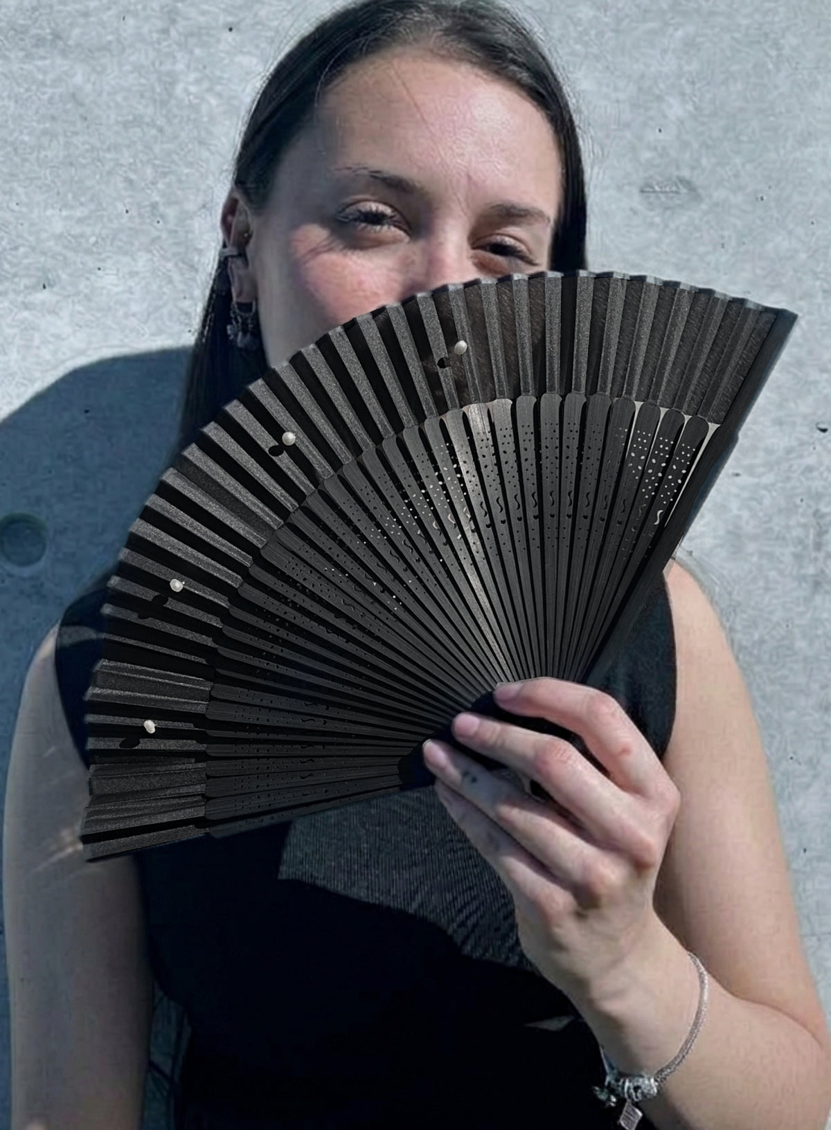 A woman holding a black folding hand fan in front of her face, wearing a black sleeveless top against a gray concrete wall.