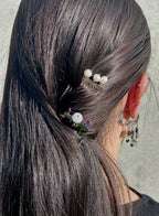 A close-up of a person's dark brown hair styled in a low ponytail, adorned with a small hair accessory featuring white pearl-like beads, green leaves, and a white flower. The person is shown from the back, wearing a silver hoop earring with dangling charms.
