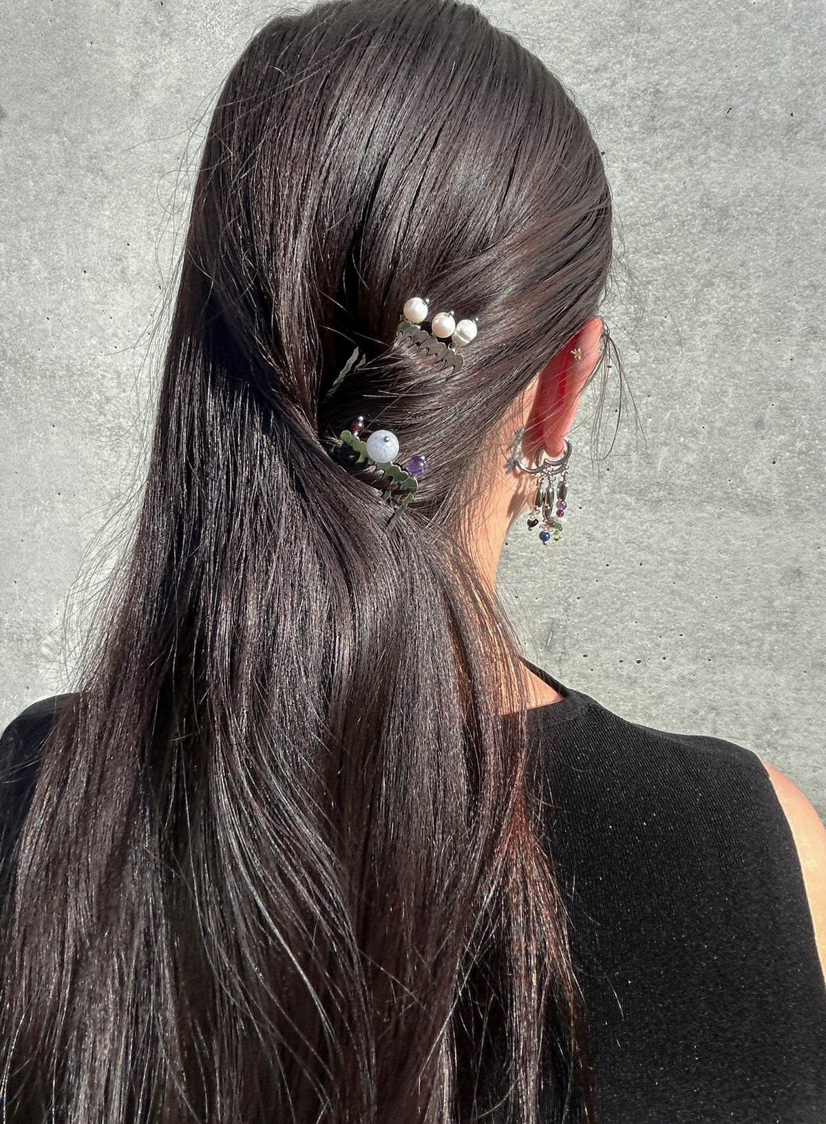 A close-up back view of a person with long, straight dark brown hair, wearing two hair accessories: a small cluster of white pearls with green leaves on one side and a floral hair clip on the other, against a textured gray wall.