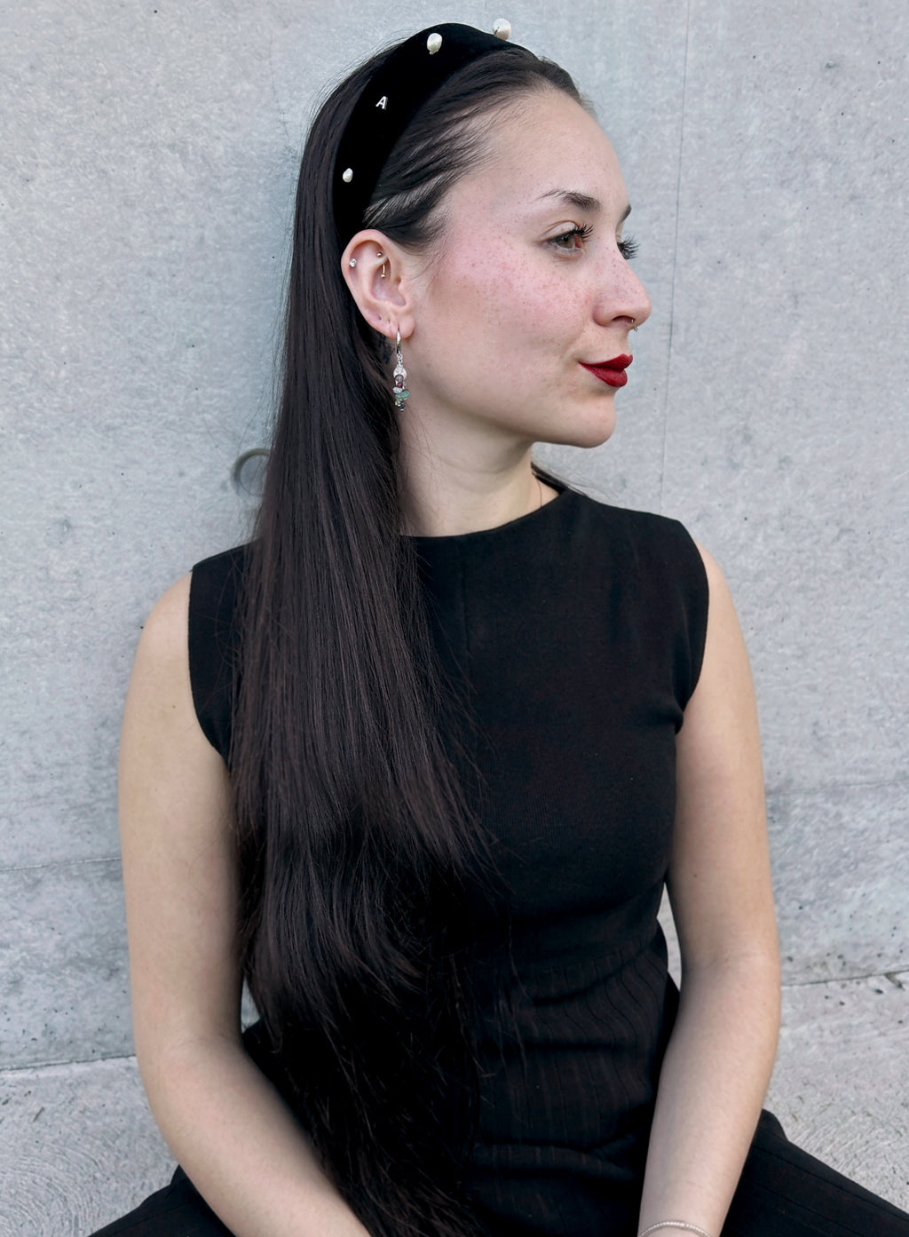 A woman with long, straight black hair is shown in profile, wearing a black fabric headband adorned with small white pearl-like beads. She is wearing a black sleeveless top and red lipstick, seated against a textured gray concrete wall.