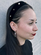 A woman in profile wearing a black velvet headband adorned with white pearl-like beads and small silver letter charms, set against a textured gray wall.
