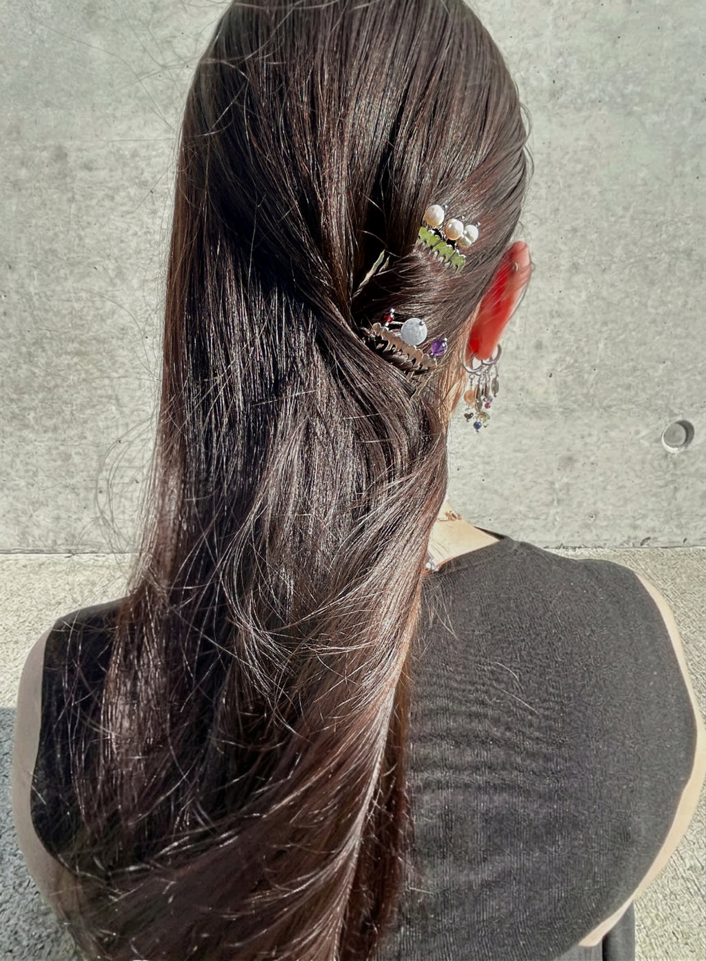A woman viewed from behind with long dark brown hair, wearing two decorative hair accessories: a pearl-and-green floral clip and a small silver-toned charm with purple and white details. She is wearing a black sleeveless top against a gray concrete wall.