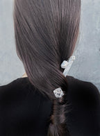 A close-up back view of a person with long, straight dark brown hair styled in a low braid, secured with two hair accessories: a white pearl-and-metal clip and a clear faceted crystal clip, against a textured gray background.