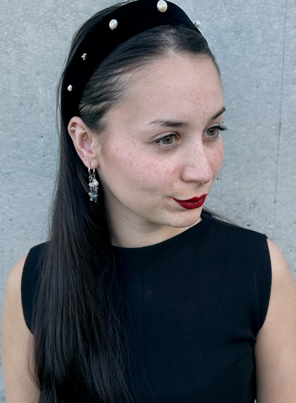 A woman with long dark hair wearing a black velvet headband adorned with white pearls and small gold studs, paired with dangling earrings and a black sleeveless top, set against a gray concrete wall.
