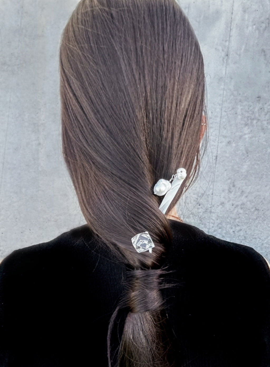 Back view of a person with long dark brown hair, wearing two silver hair accessories: a larger barrette with pearl-like beads and a smaller clear faceted hair clip, against a gray textured background.