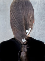Back view of a person with long dark brown hair, wearing two silver hair accessories: a larger barrette with pearl-like beads and a smaller clear faceted hair clip, against a gray textured background.