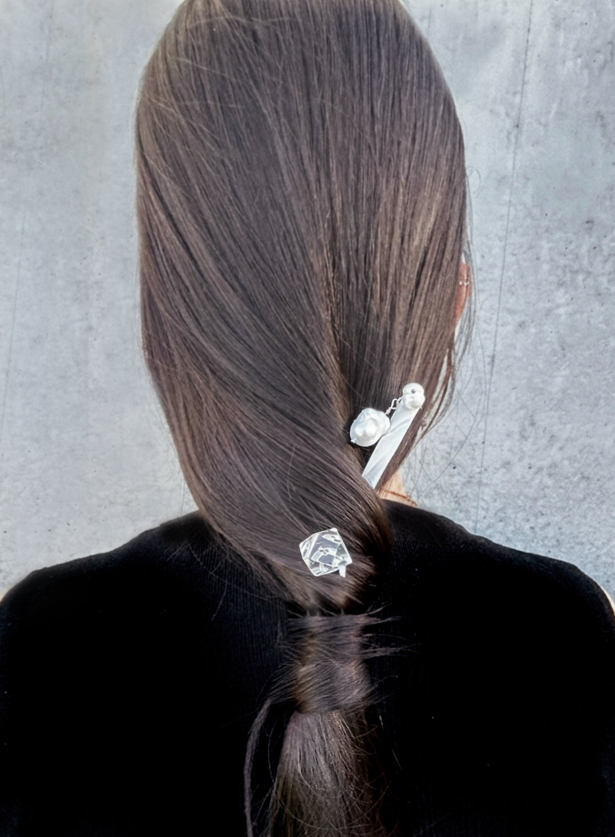 Back view of a person with long dark brown hair, wearing two silver hair accessories: a larger barrette with pearl-like beads and a smaller clear faceted hair clip, against a gray textured background.
