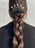 A close-up back view of a person with long brown hair in a thick braid, secured with a black hair claw clip adorned with gold pins and white pearl-like beads, against a light gray wall.