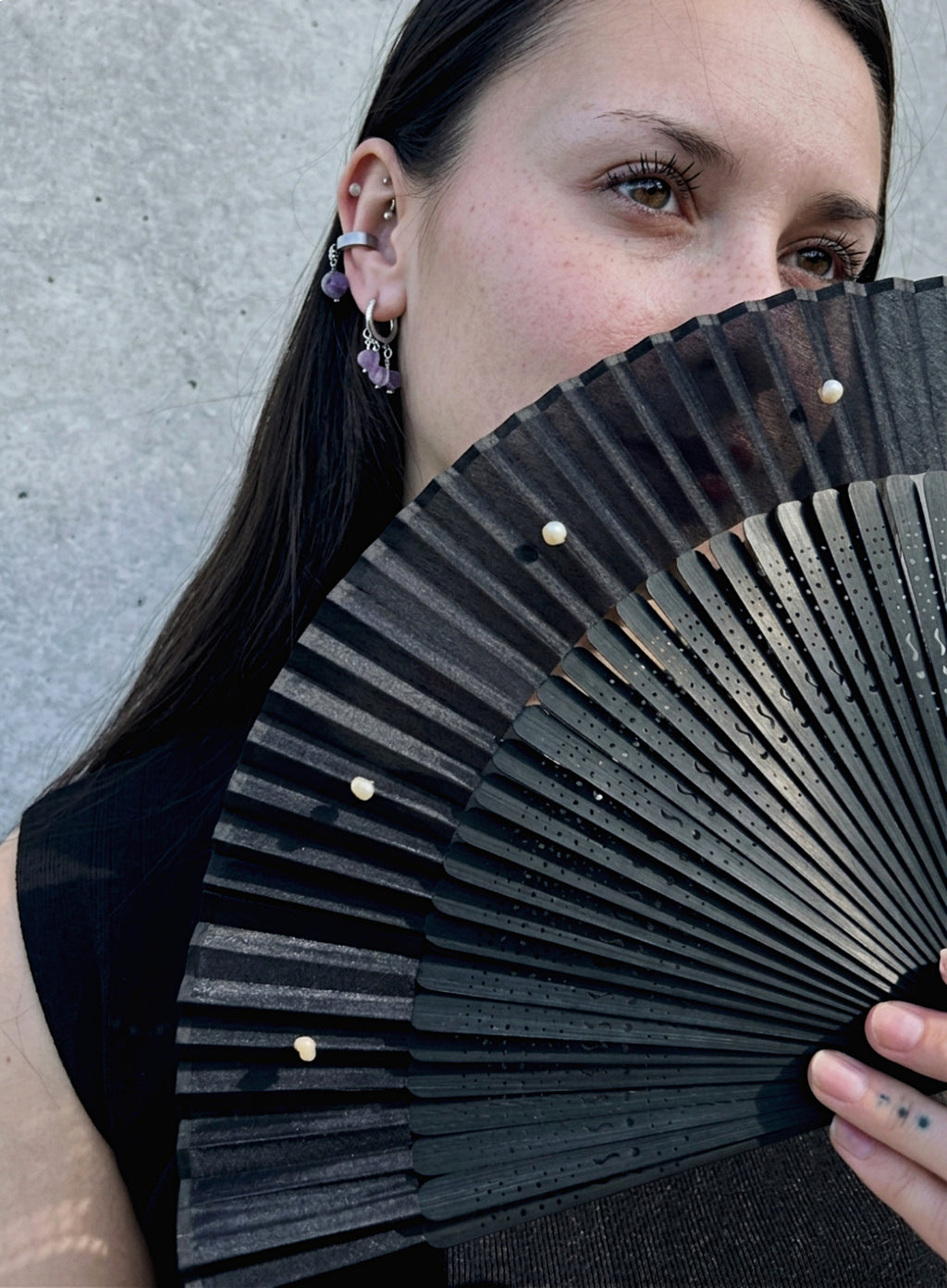A close-up lifestyle shot of a woman holding a black folding hand fan with small white pearls. She is wearing multiple piercings, including a purple beaded hoop earring, against a neutral gray concrete wall.