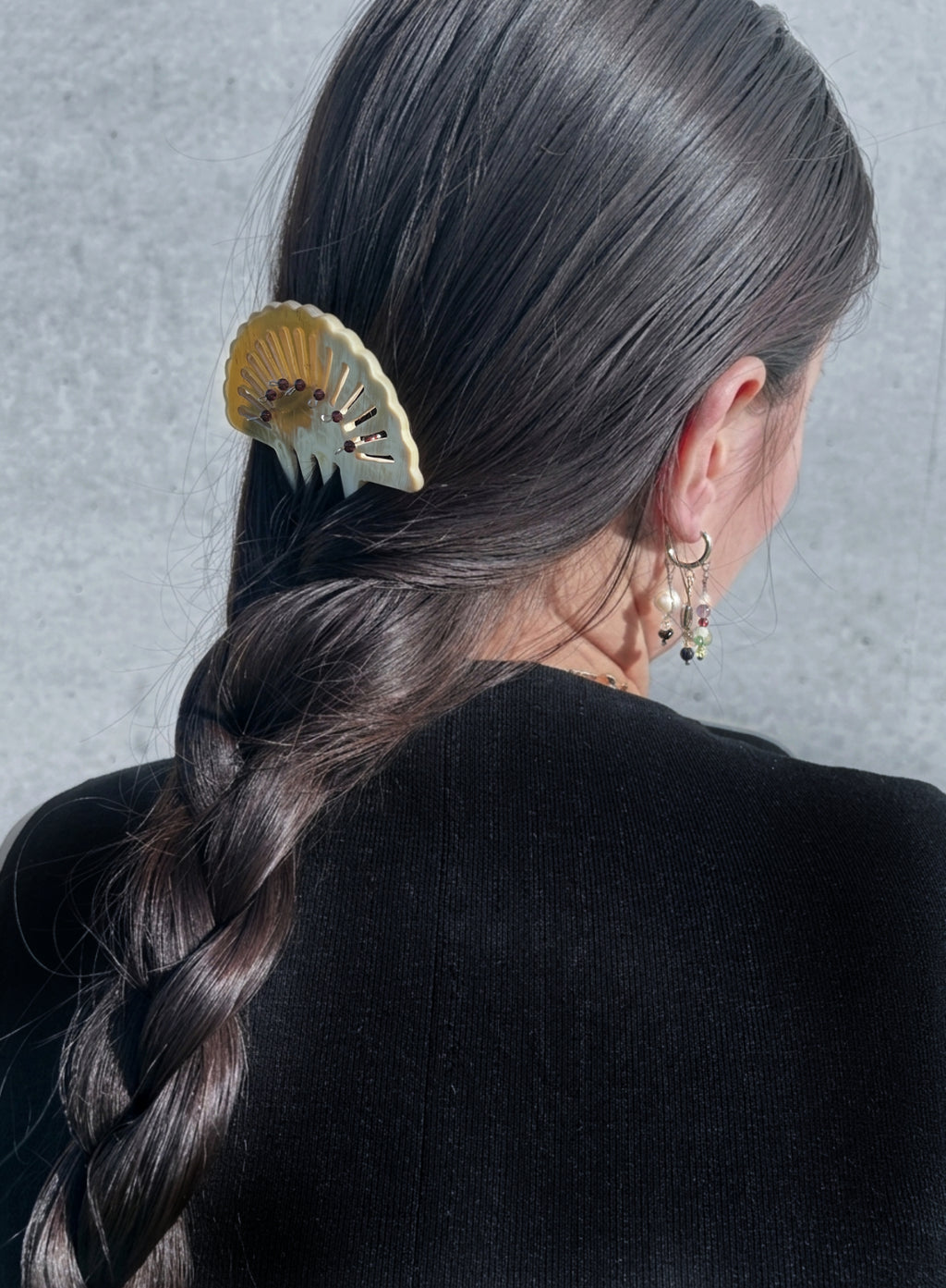 A close-up back view of a person with long dark brown hair in a side braid, wearing a cream and gold fan-shaped hair claw clip. The person is also wearing small gold hoop earrings with dangling charms, set against a textured gray wall.