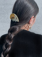 A close-up back view of a person with long dark brown hair in a side braid, wearing a cream and gold fan-shaped hair claw clip. The person is also wearing small gold hoop earrings with dangling charms, set against a textured gray wall.