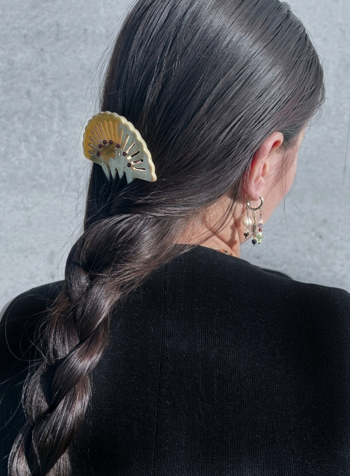 A close-up back view of a person with long dark brown hair in a side braid, wearing a cream and gold fan-shaped hair claw clip. The person is also wearing small gold hoop earrings with dangling charms, set against a textured gray wall.