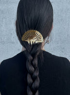 A close-up back view of a person with long black hair in a thick braid, secured with a metallic gold fan-shaped hair claw clip against a textured gray wall.