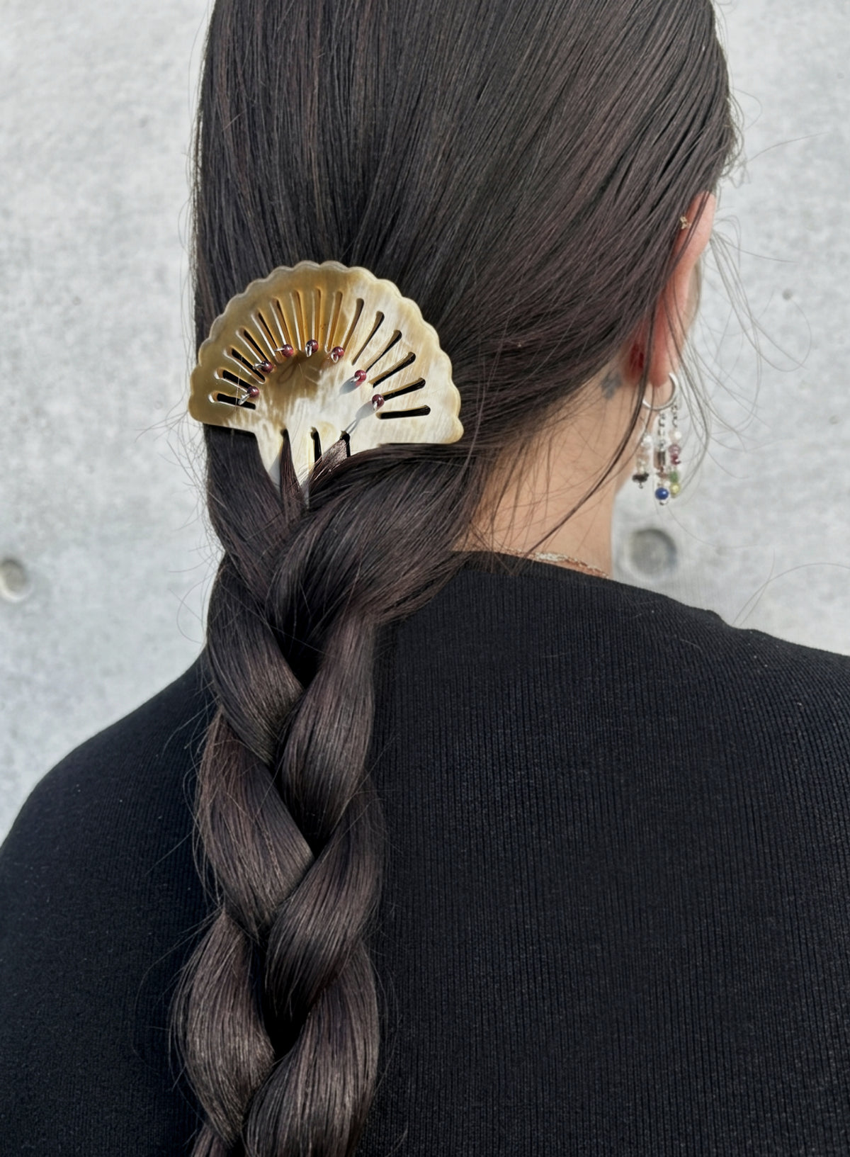 A close-up lifestyle shot of a woman from behind, showcasing a decorative gold fan-shaped hair claw clip holding her dark brown braided hair. She is wearing a black top and dangling earrings against a textured gray concrete wall.