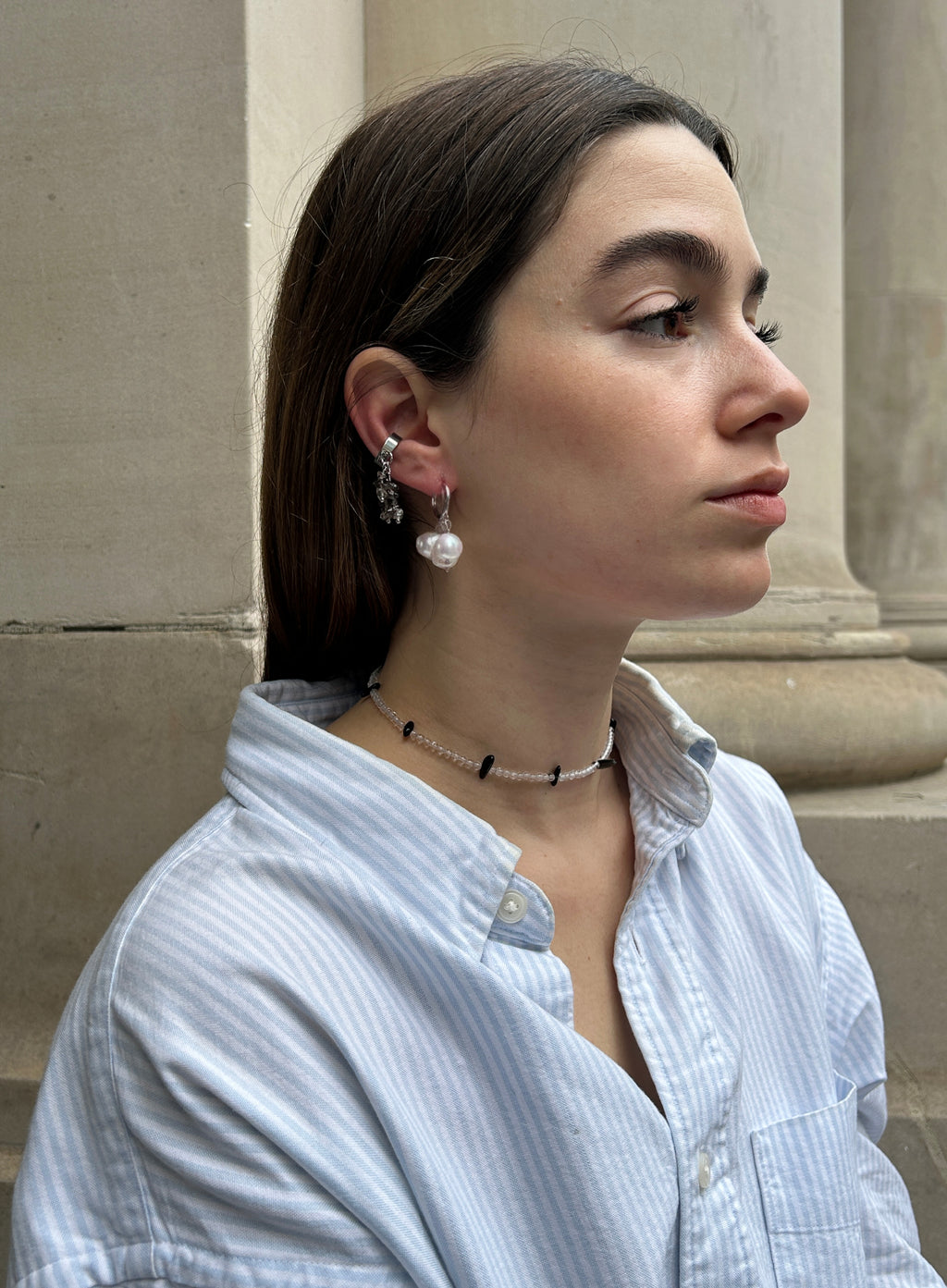A woman in a light blue and white striped button-down shirt is shown in profile, wearing a delicate black beaded choker necklace and a dangling silver earring with a white pearl.