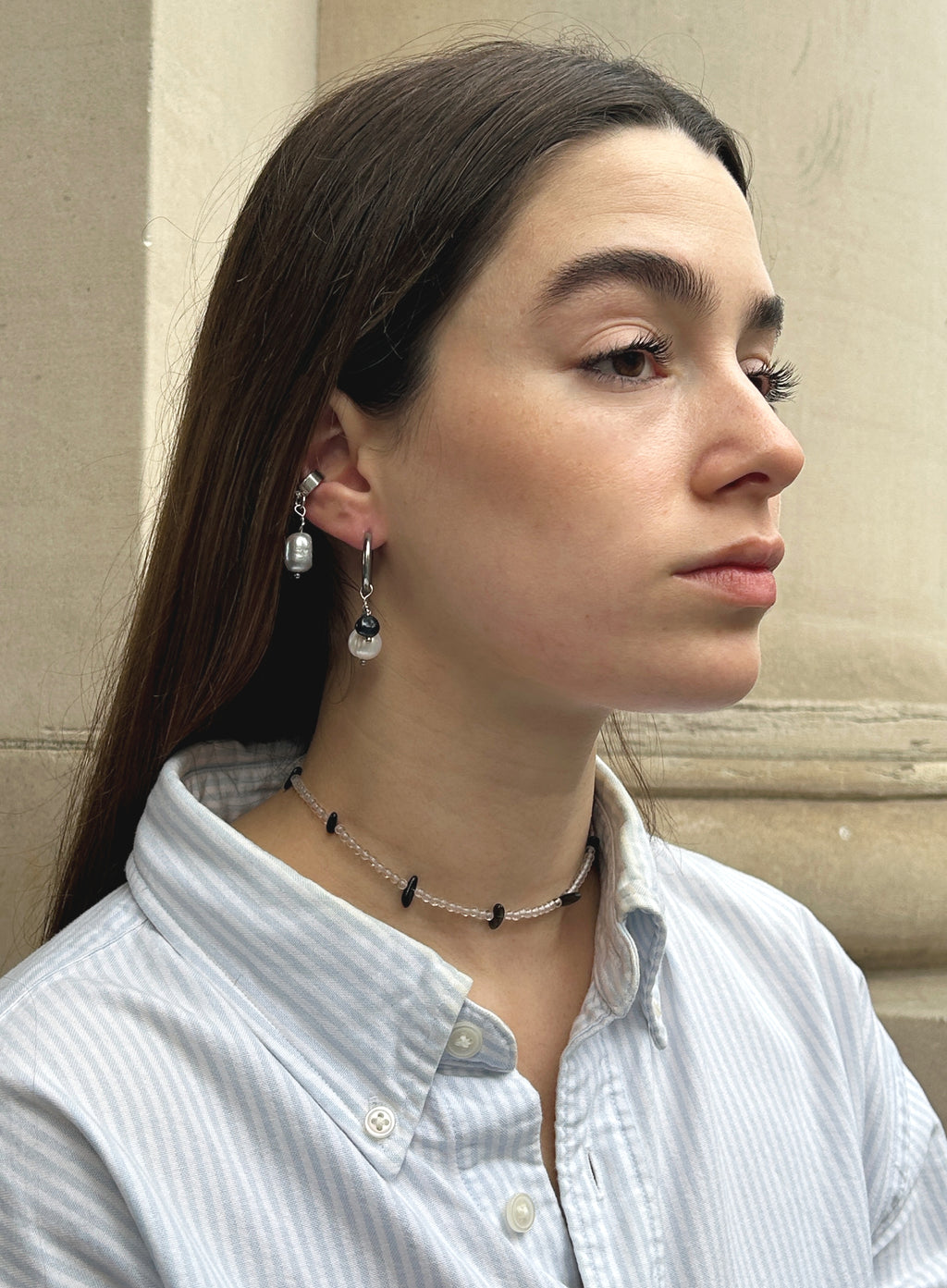 A woman in a light blue and white striped button-down shirt is shown in profile, wearing silver-toned drop earrings with clear beads and a matching beaded choker necklace.