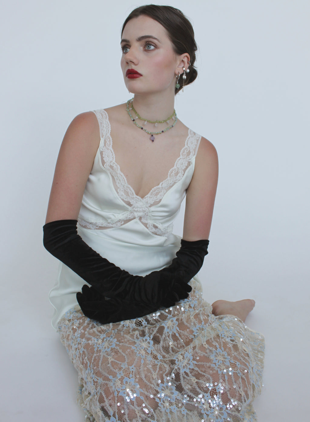 A woman seated against a plain light gray studio background, wearing a white satin camisole with lace trim, long black velvet opera gloves, layered necklaces, and a sheer silver sequined skirt.