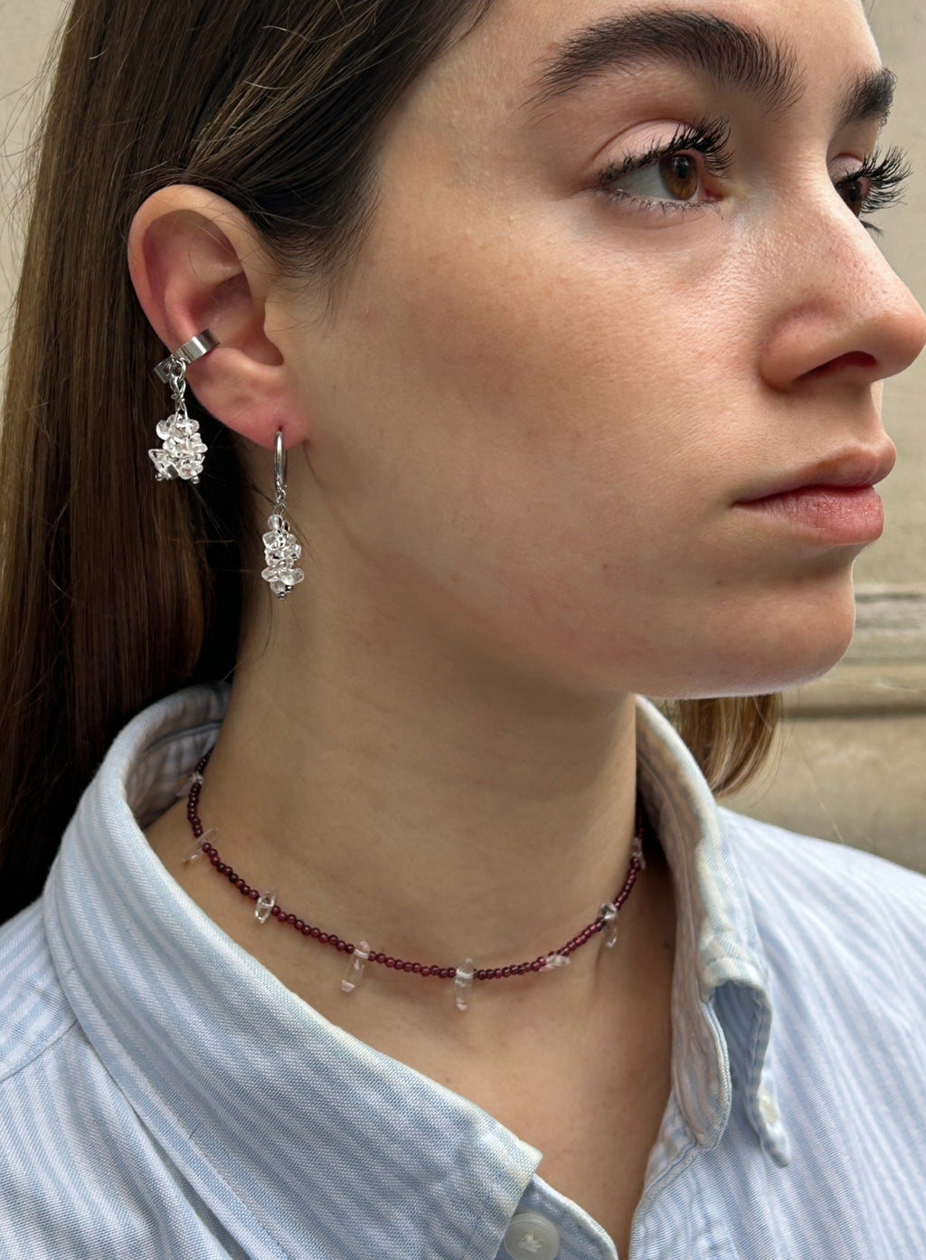 A close-up side profile of a woman wearing a silver-toned dangle earring with a floral cluster drop, a matching beaded choker necklace, and a light blue striped button-up shirt.