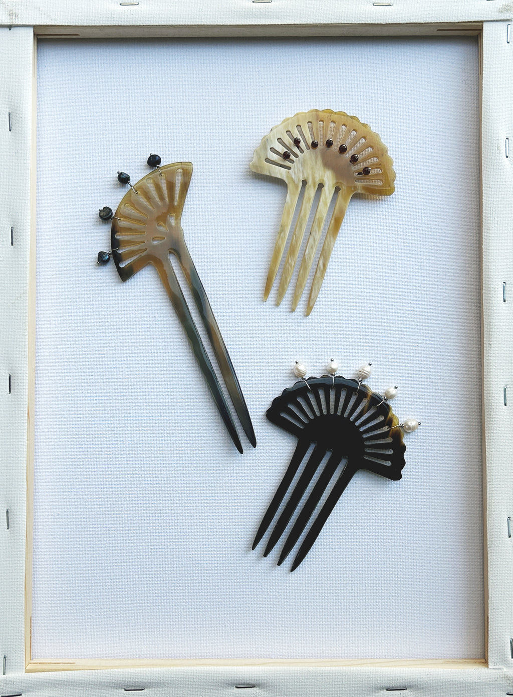 Three decorative hair combs arranged on a white background inside a light wood frame. The combs have fan-like cutouts and are shown in black, beige, and brown tones.