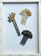 Three decorative hair combs arranged on a white background inside a light wood frame. The combs have fan-like cutouts and are shown in black, beige, and brown tones.