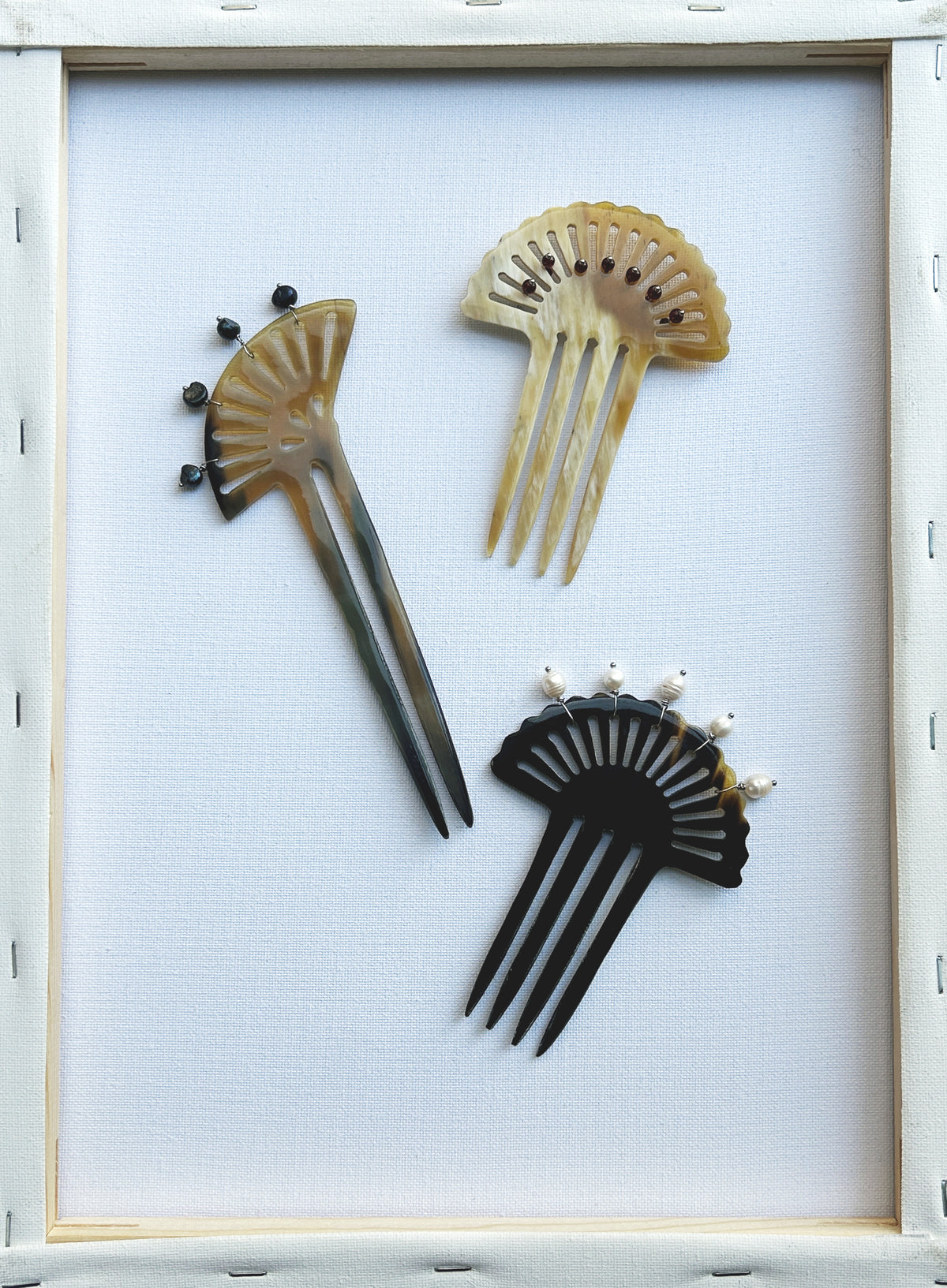 Three decorative hair combs arranged on a white background inside a light wood frame. The combs have fan-like cutouts and are shown in black, beige, and brown tones.