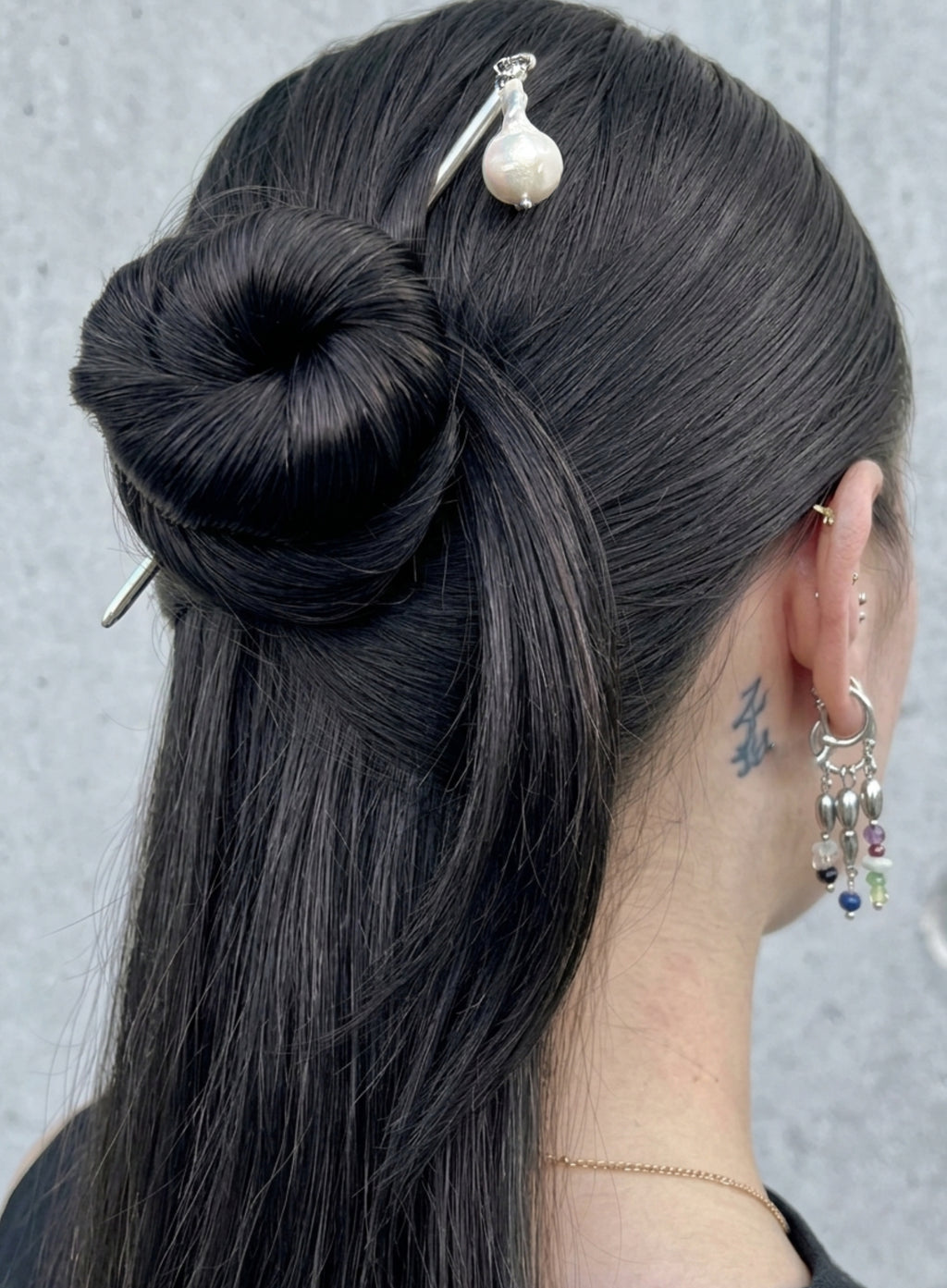 A close-up back view of a person with long, straight black hair styled in a low bun, accessorized with a silver hair stick featuring a pearl top and a silver earring with dangling colorful beads.