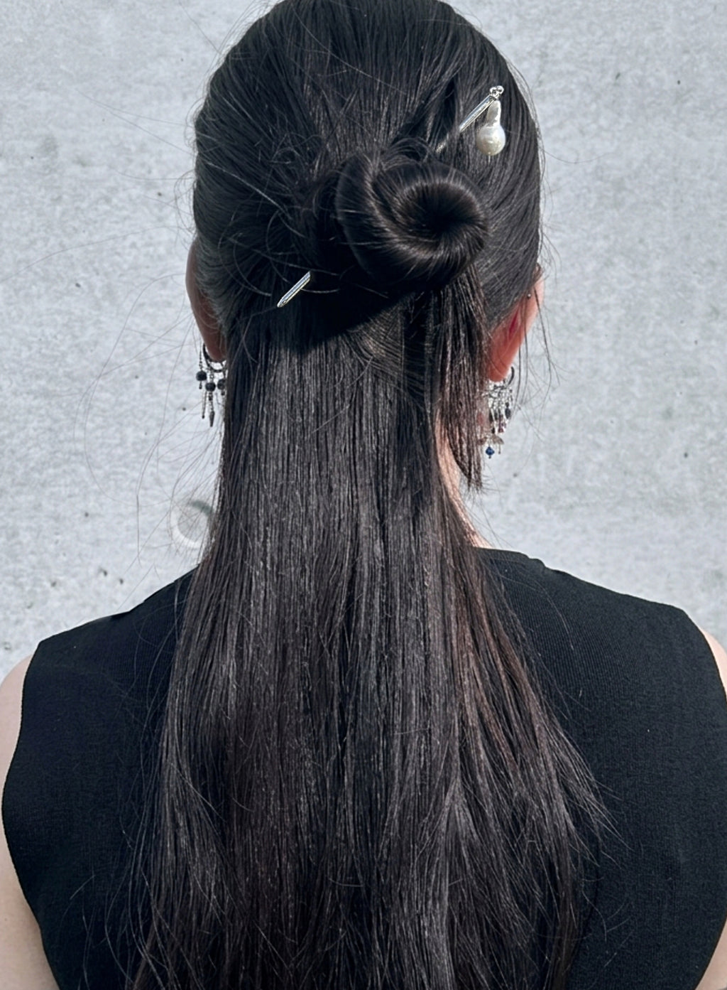 A person shown from the back wearing a black sleeveless top, with long straight black hair styled in a half-up bun secured with a silver hair clip and pearl hairpin. The background is a textured light gray wall.