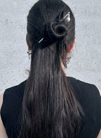A person shown from the back wearing a black sleeveless top, with long straight black hair styled in a half-up bun secured with a silver hair clip and pearl hairpin. The background is a textured light gray wall.