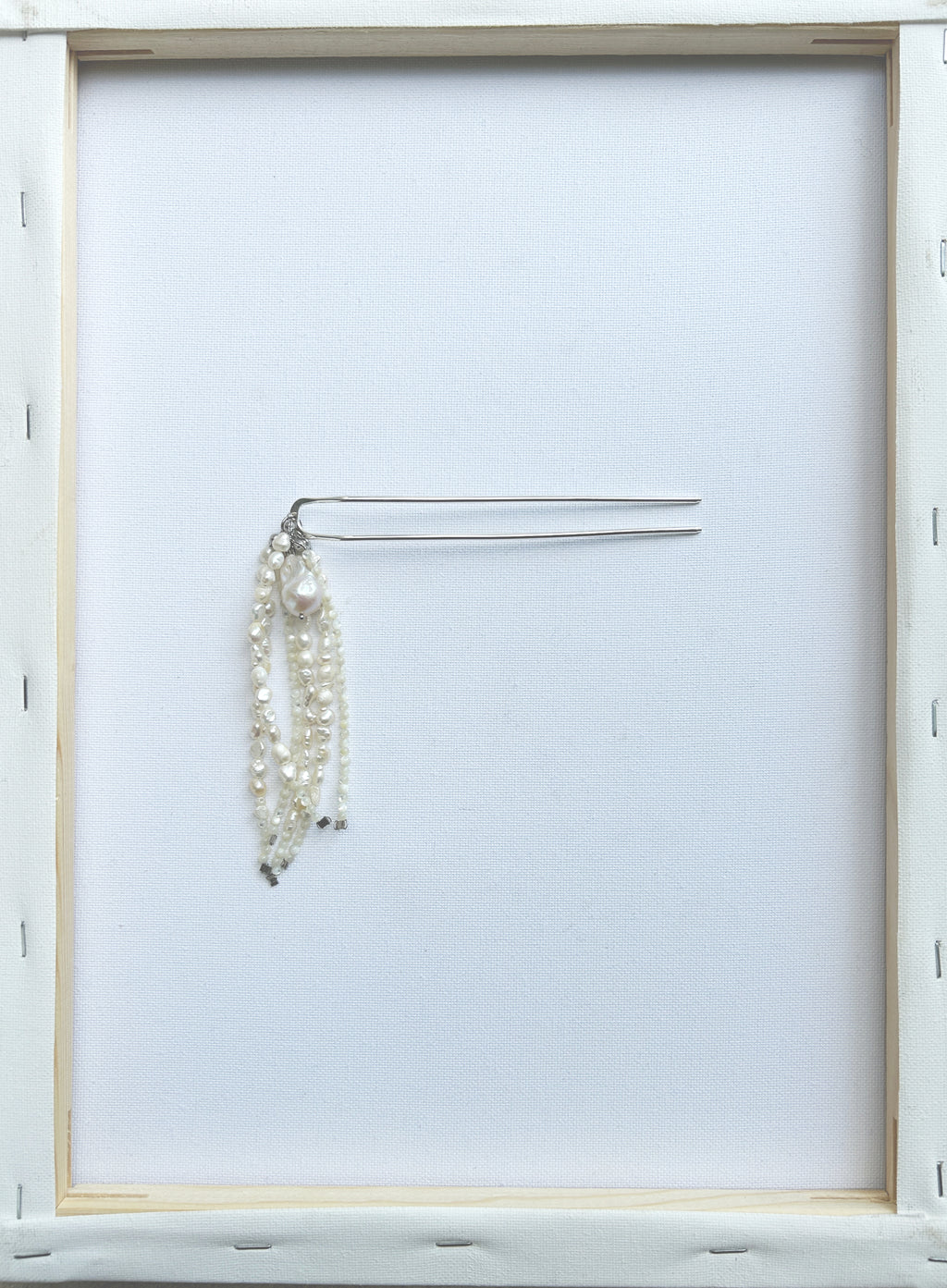 A silver-tone metal hair comb with three dangling strands of white faux pearls, displayed on a clean white background inside a light wood frame.