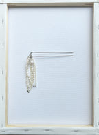 A silver-tone metal hair comb with three dangling strands of white faux pearls, displayed on a clean white background inside a light wood frame.