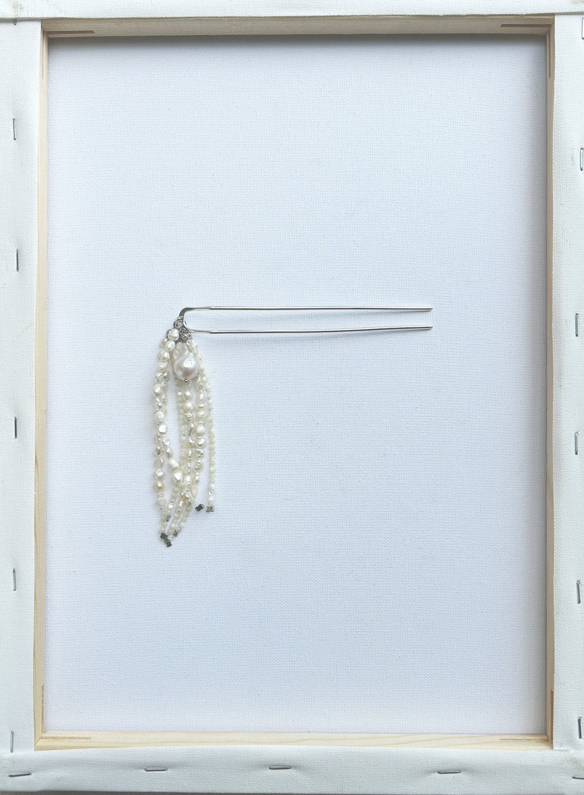 A silver-tone metal hair comb with three dangling strands of white faux pearls, displayed on a clean white background inside a light wood frame.